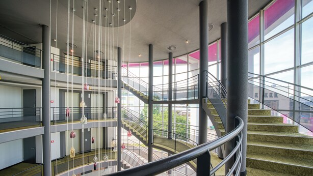 Ein mehrgeschossiges Interieur mit einer zentralen Treppe, Glasgeländern und Blick ins Freie.