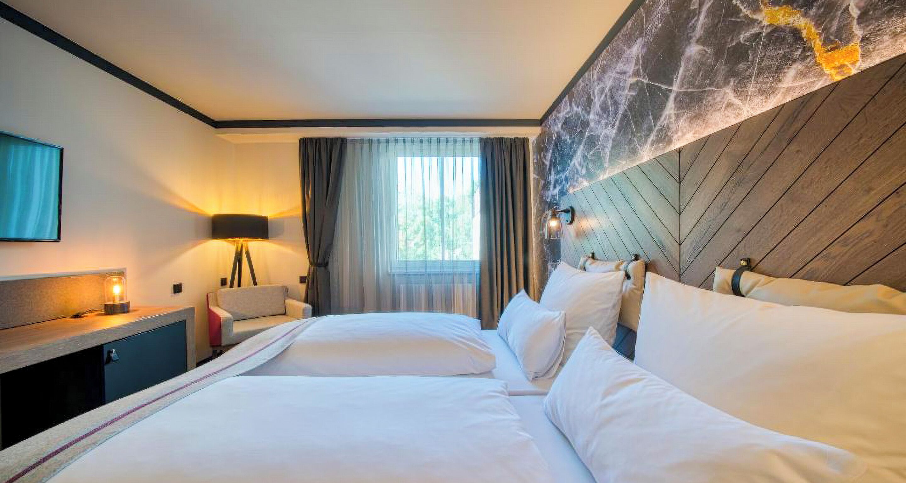 A hotel room with a bed, bedside table, lamp, and a view of the outdoors.