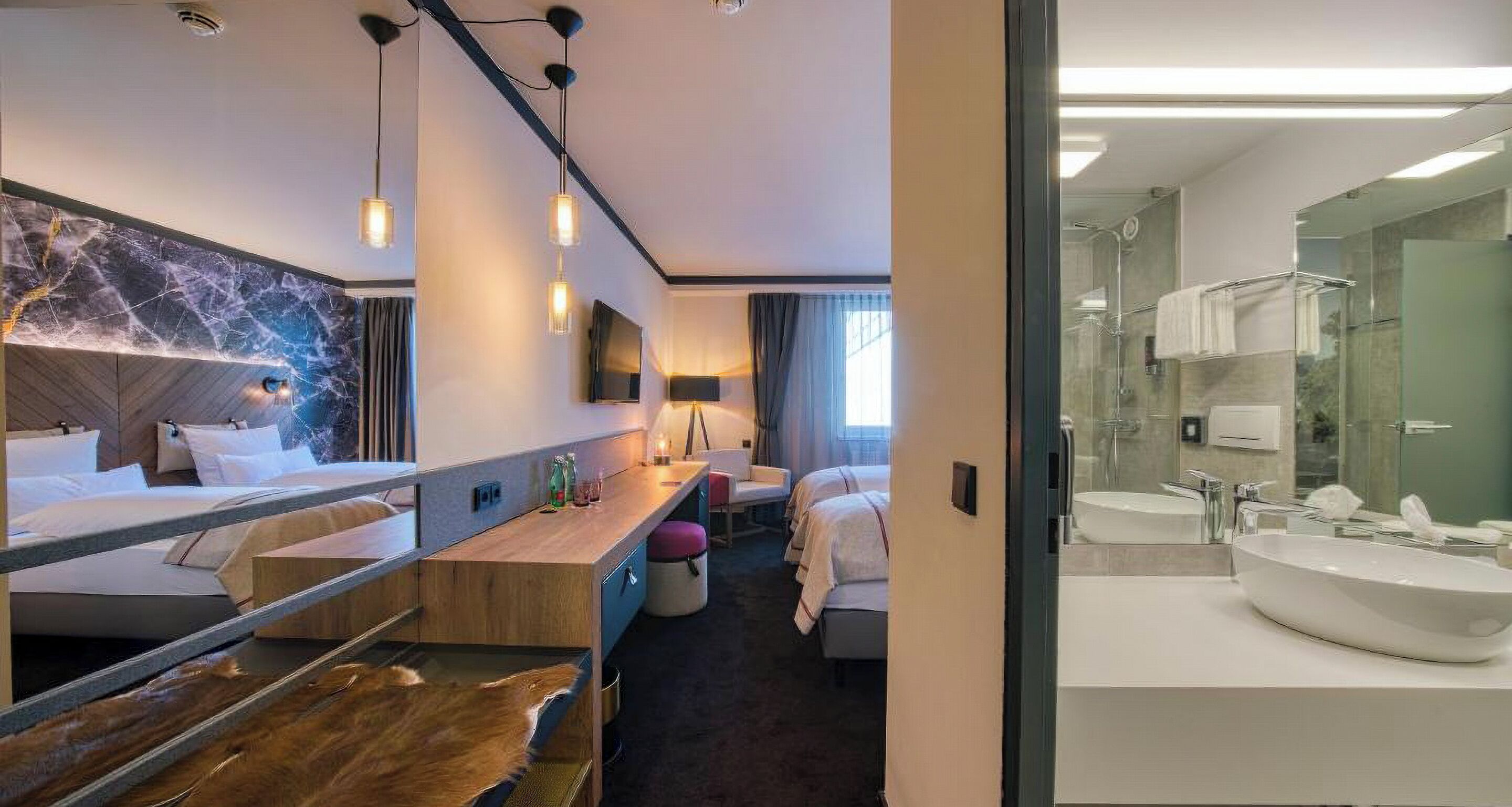 Habitación de hotel con cama, escritorio, silla, espejo y baño con lavabo y ducha.