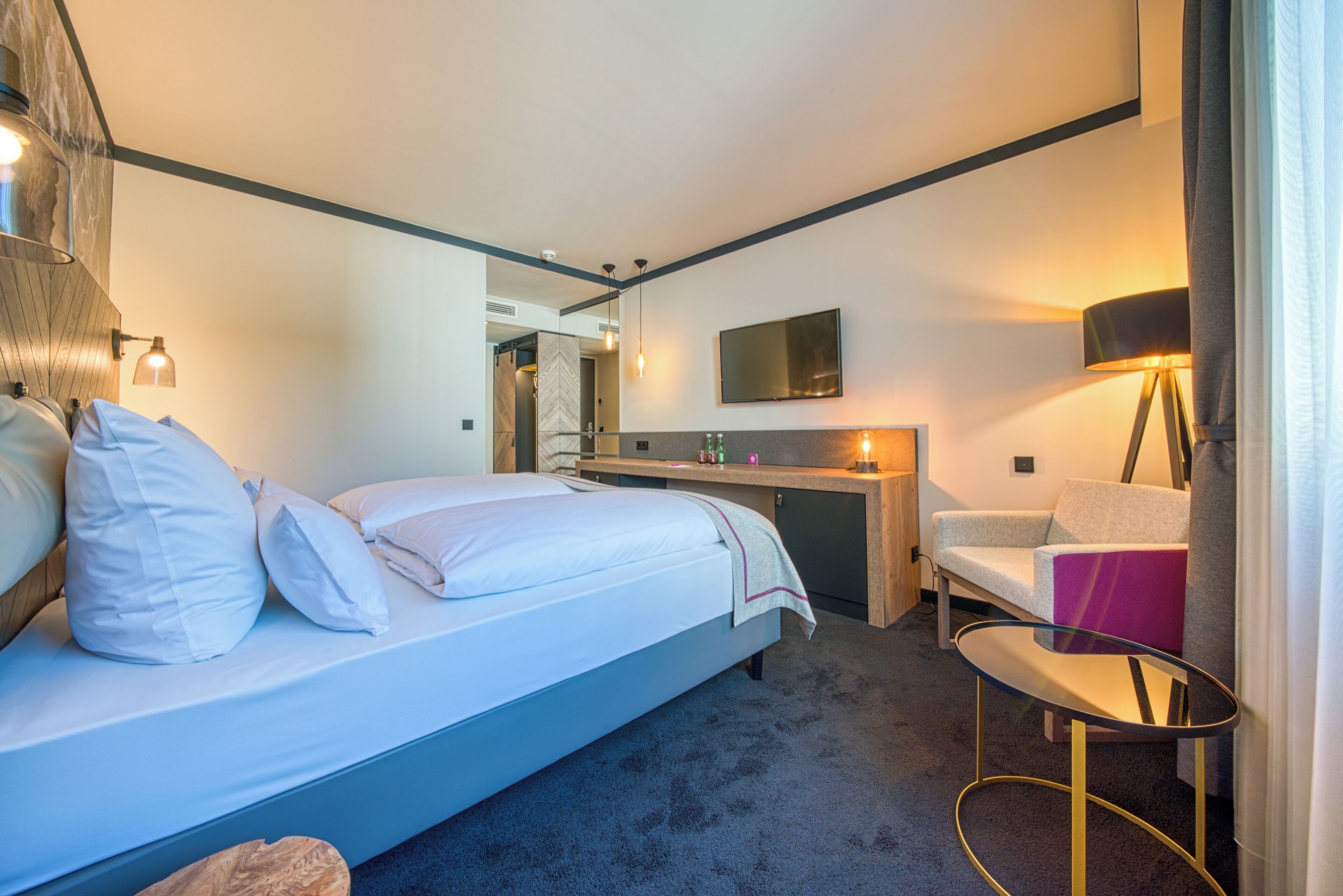 Ein Hotelzimmer mit Bett, Nachttisch, Schreibtisch, Stuhl und einem an der Wand befestigten Fernseher.