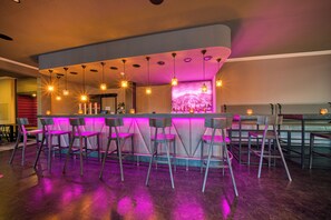 A bar area with a curved counter, multiple bar stools, pendant lighting, and a large screen displaying a mountain scene. - PLAZA INN Salzburg Messe  (Salzburg)