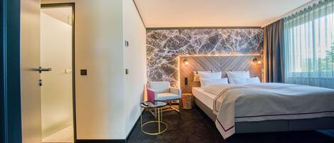 Habitación de hotel con una cama grande, zona de estar con una silla y una mesa, pared decorativa de mármol y vistas al exterior.