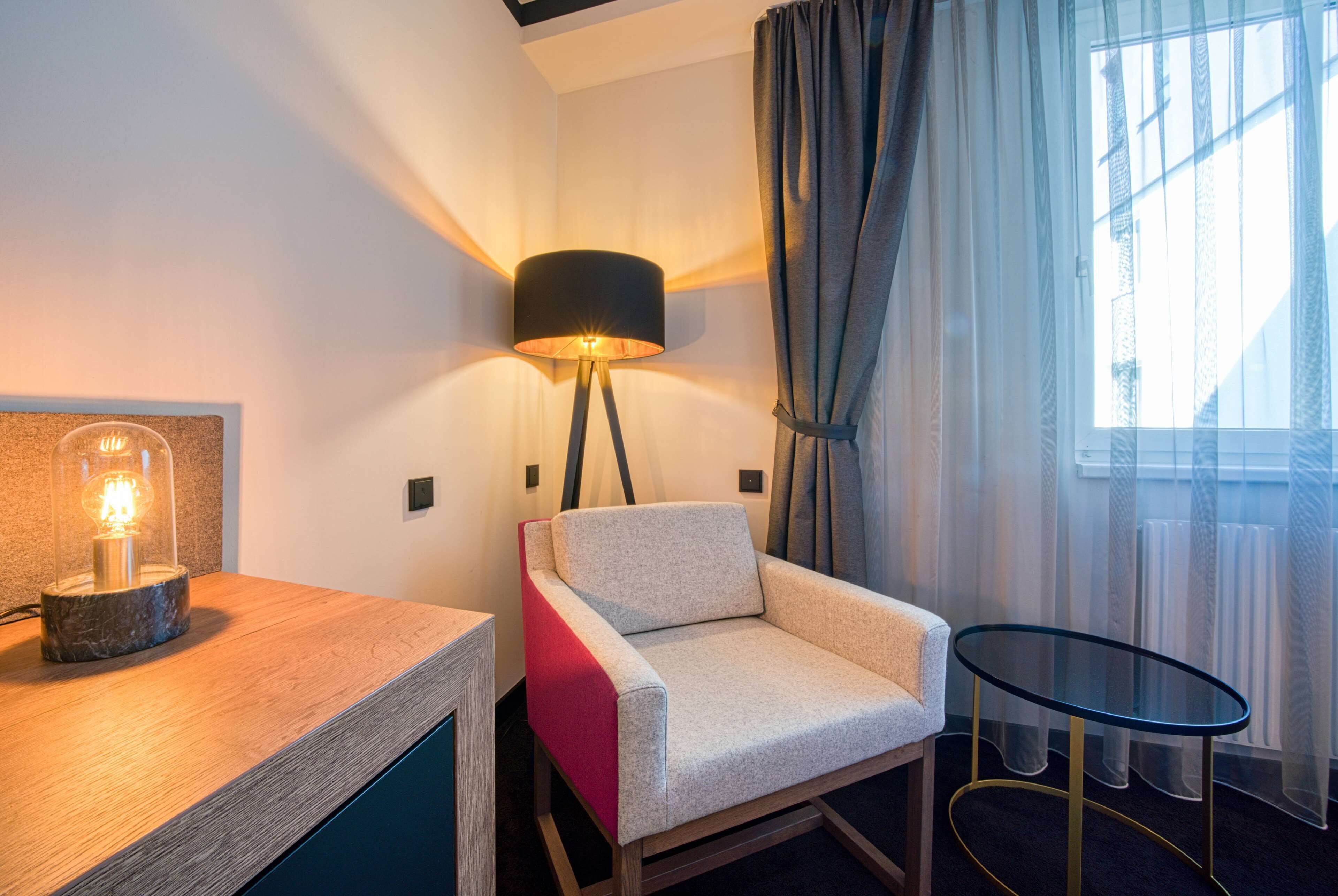A modern hotel room with a wooden side table, a lamp, a cushioned armchair, a glass coffee table, and a view of the outdoors through sheer curtains.