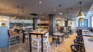 A modern restaurant interior with multiple dining areas, a kitchen in the background, and pendant lighting. - PLAZA INN Salzburg Messe  (Salzburg)
