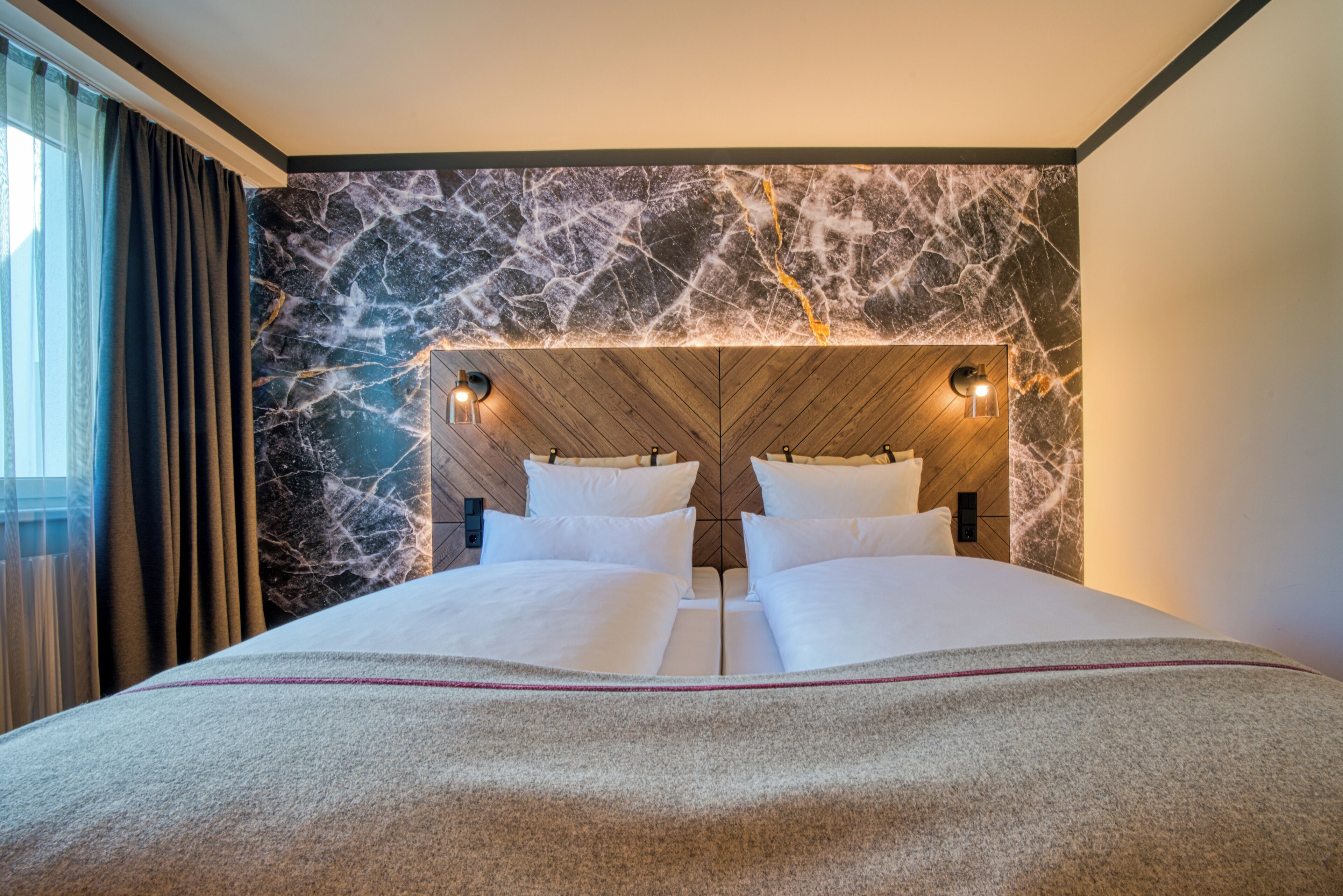 Habitación de hotel con una cama grande, cabecero de madera, pared de mármol y dos luces fijadas a la pared.