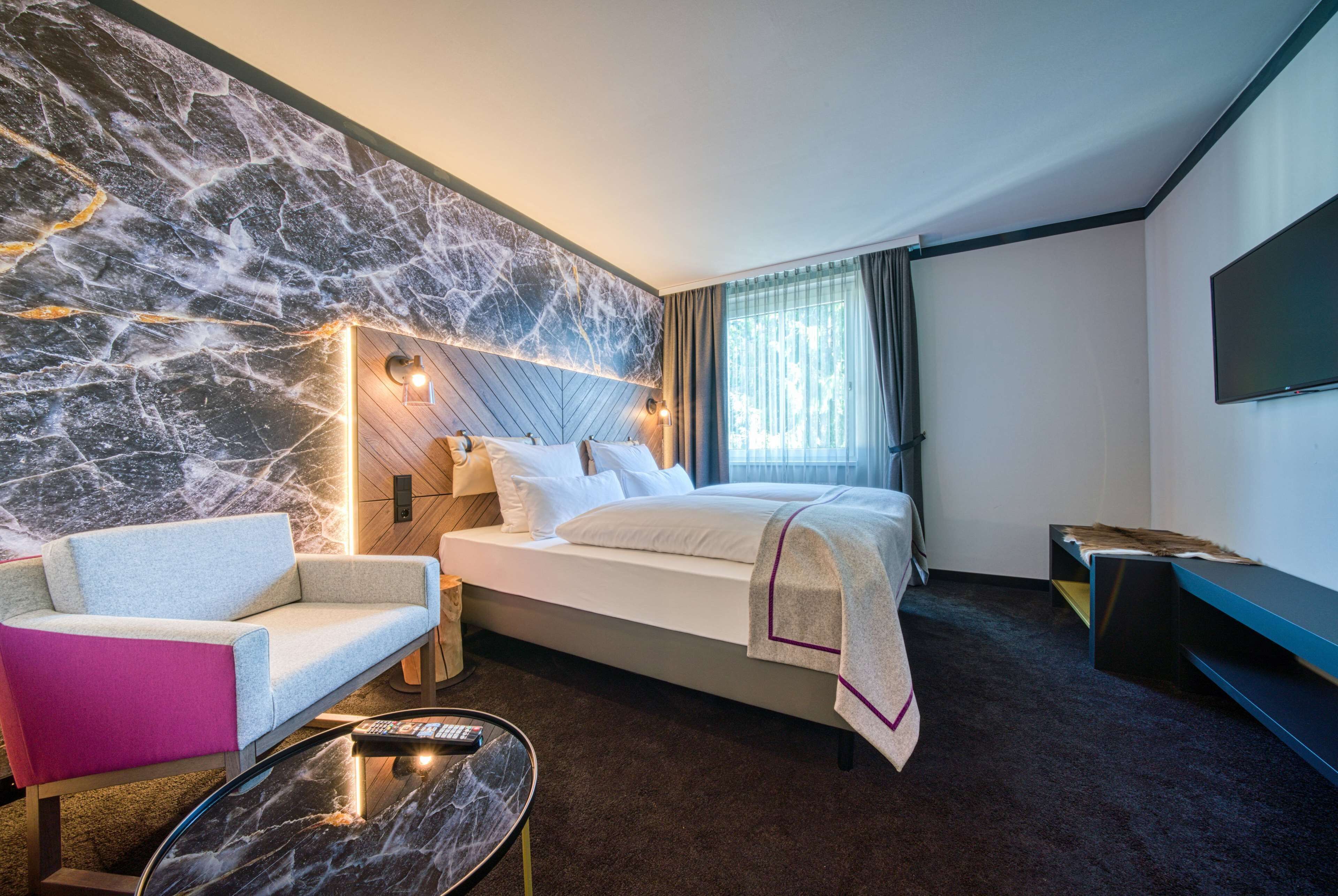 A modern hotel room with a marble accent wall, a bed with a headboard, a sofa, a TV, and a small table.