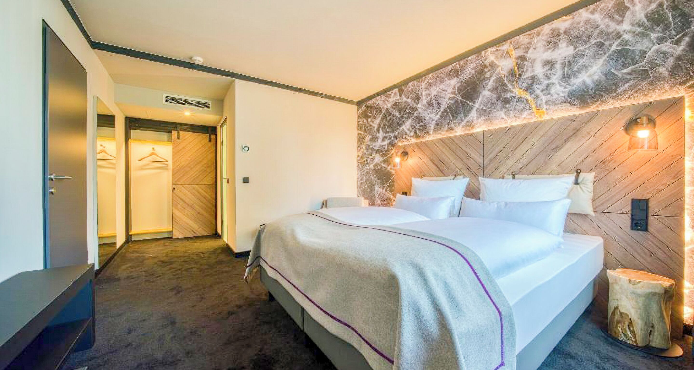 A modern hotel room with a large bed, a wooden headboard, and a stone accent wall.