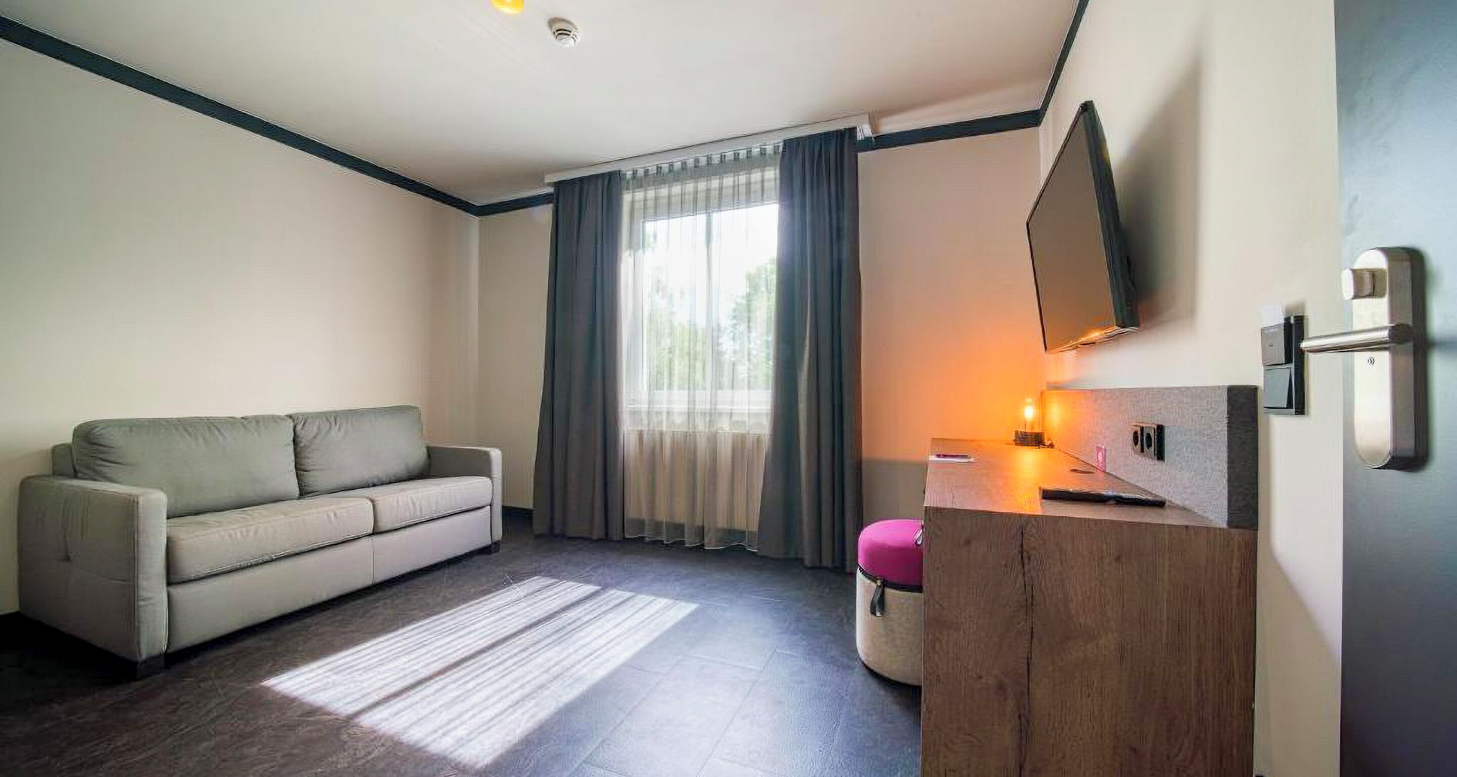 A modern hotel room with a sofa, a wooden desk, a flat-screen TV, and a window with curtains.