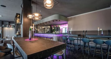 Un'area bar moderna con un bancone in marmo, sgabelli da bar, illuminazione a sospensione e un bancone bar illuminato di viola.