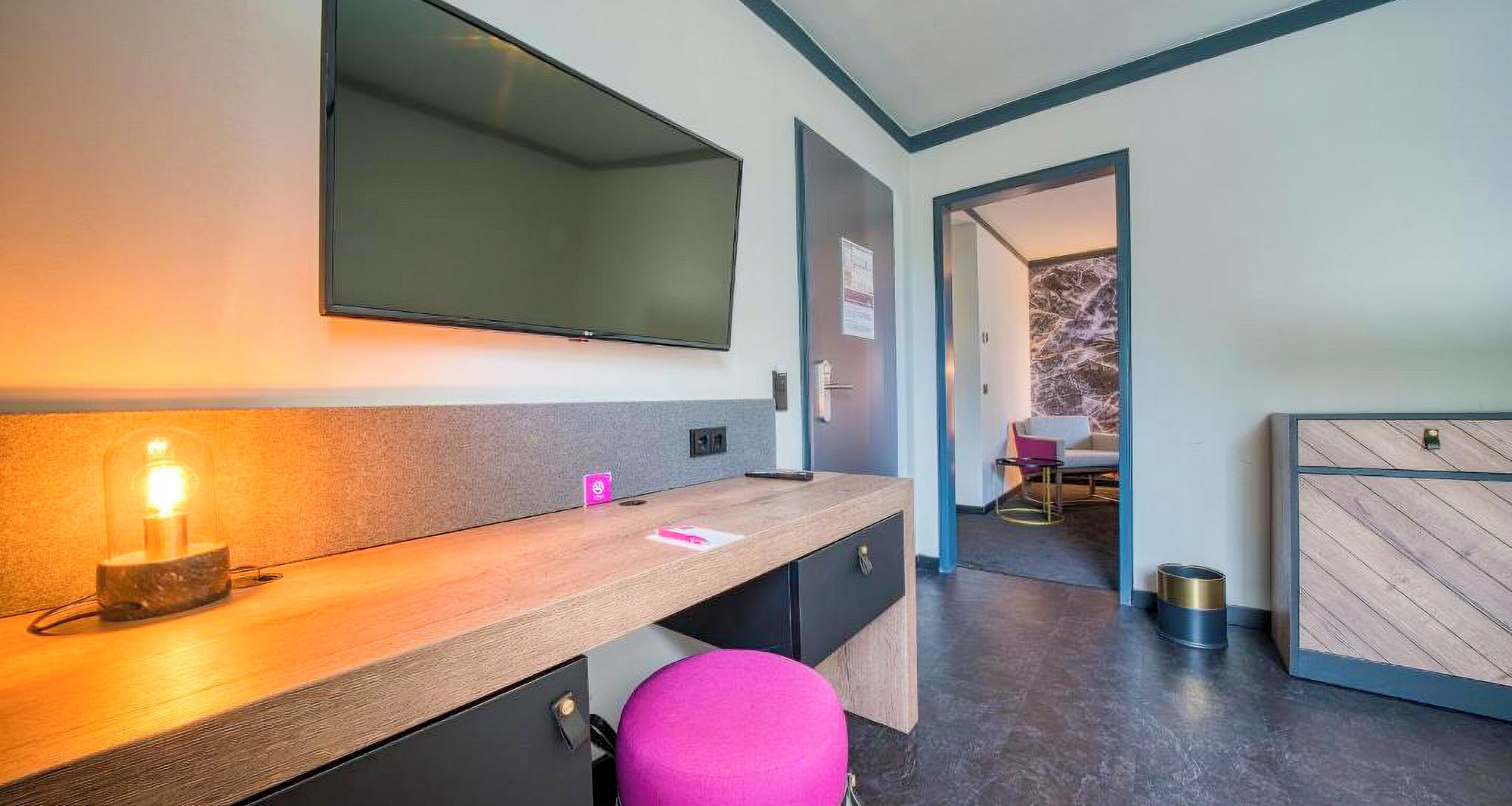 A modern bathroom with a wooden vanity, a mirror, a pink stool, and a visible shower area.