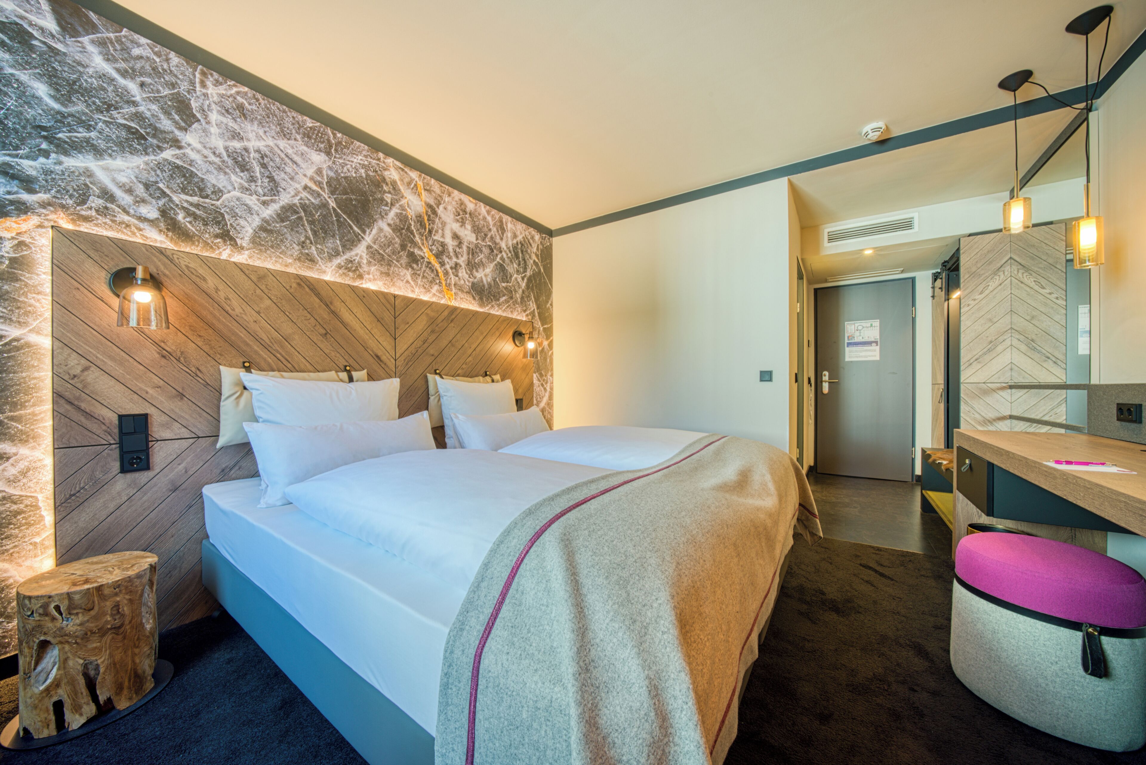 Una habitación de hotel moderna con cabecera de madera, una cama con sábanas blancas, un taburete de madera pequeño, un banco y un espejo en la pared.