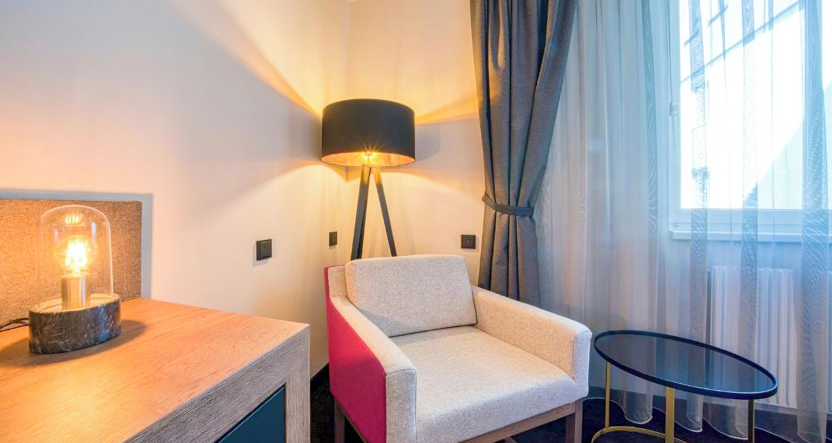 A hotel room with a modern lamp, a chair, a small table, and a view of the outside through sheer curtains.