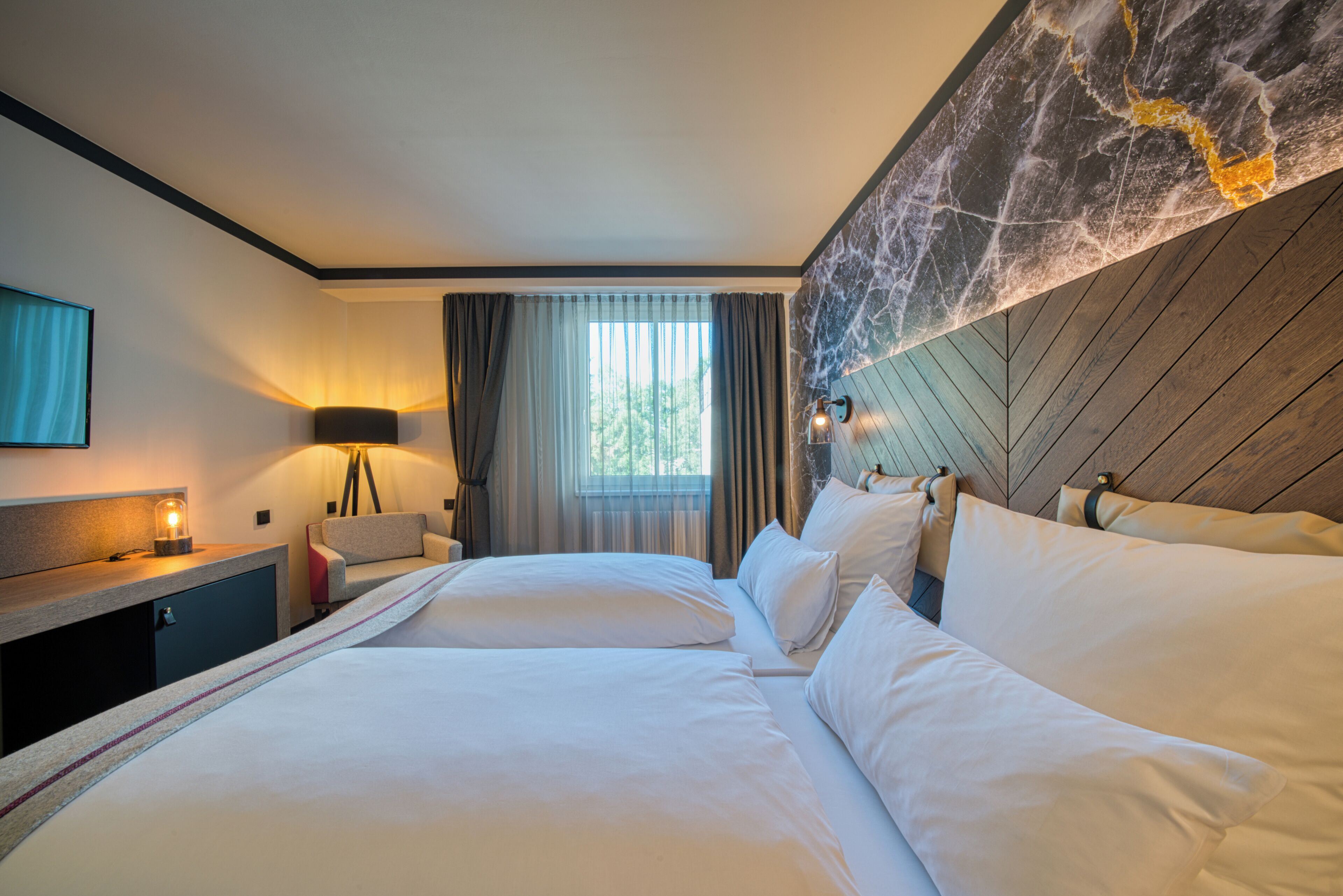 A hotel room with a large bed, a desk with a lamp, a window with curtains, and a marble accent wall.