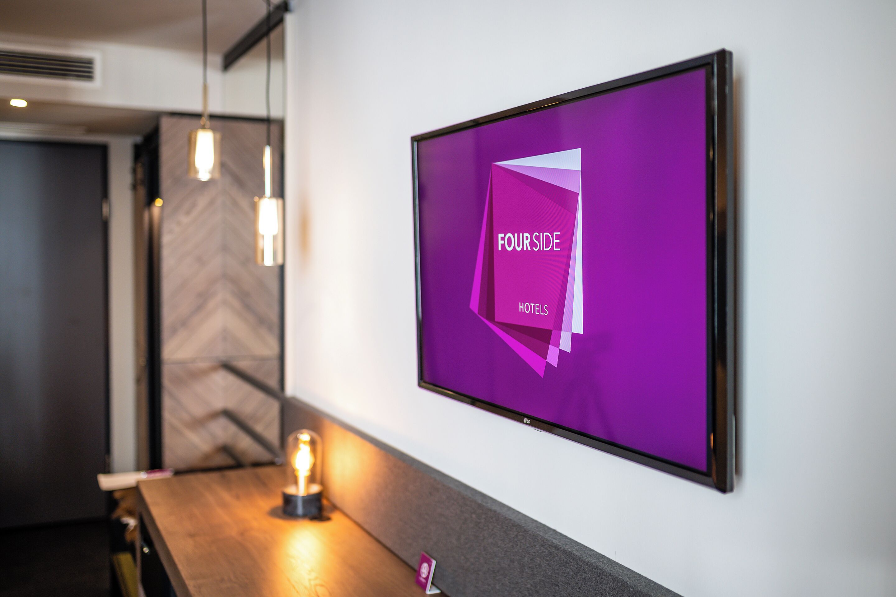 Un interior de hotel moderno con una pantalla grande que muestra 'FOURSIDE HOTELS' y un mostrador de recepción de madera con iluminación colgante.