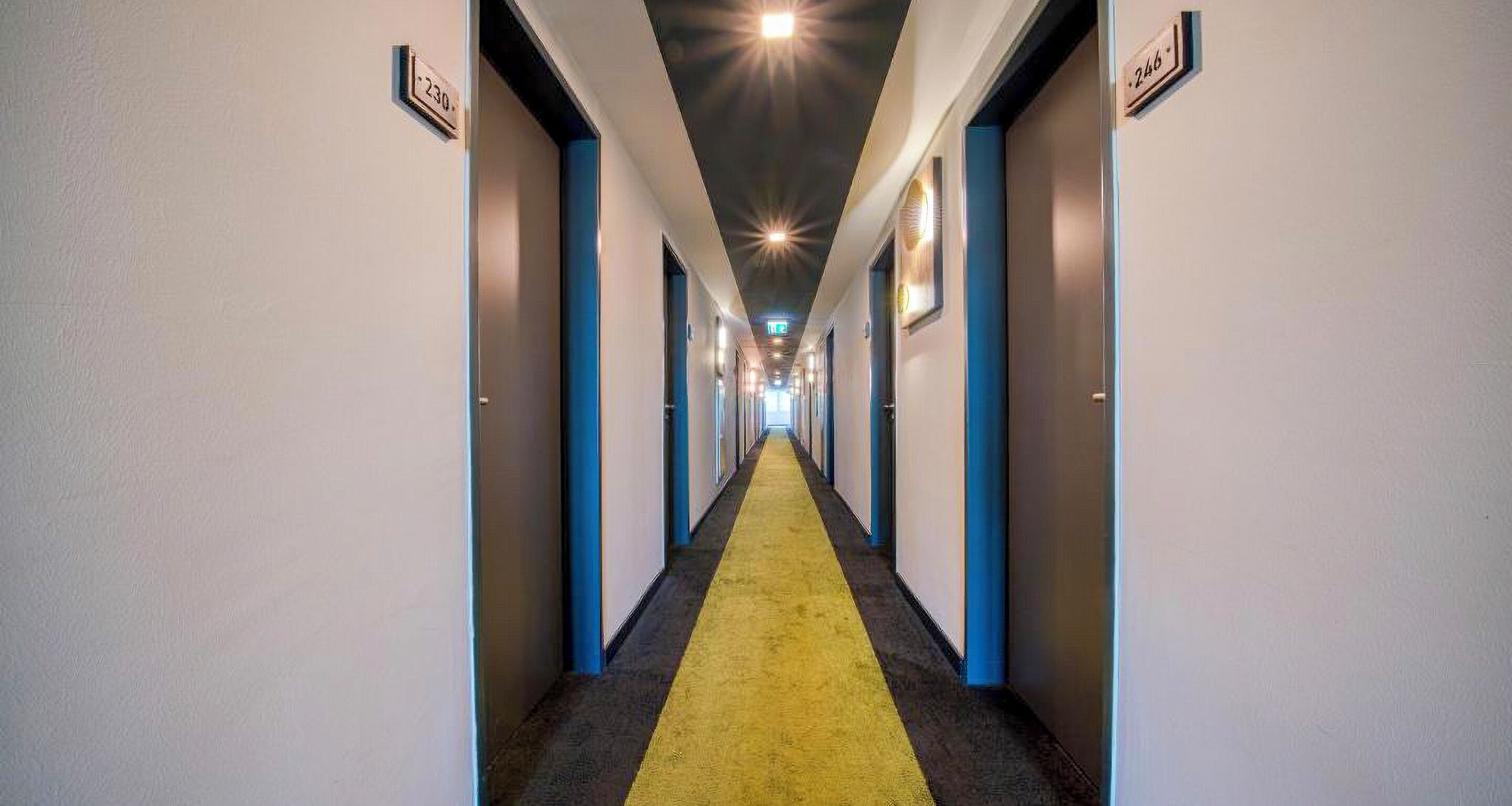 A hotel corridor with a yellow carpet, blue doors, and numbered room signs.