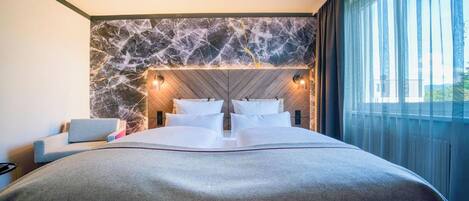Quarto de hotel moderno com cabeceira estampada em mármore, uma cama grande com lençóis brancos, uma mesinha de cabeceira e vista para o exterior através de cortinas finas.