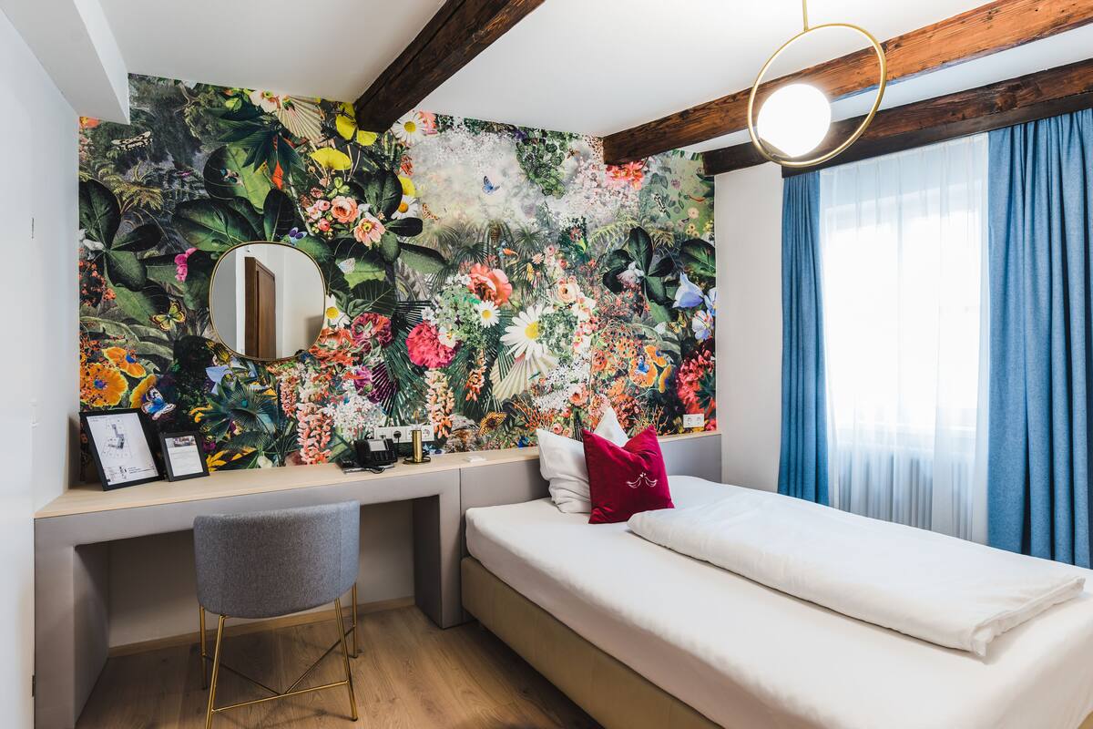 Un dormitori amb un llit, un escriptori, una cadira, un mirall i una paret decorada amb un mural floral.