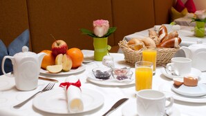 A breakfast setting with a table set for two, including plates, cutlery, a teapot, a basket of bread, and a vase with a flower.