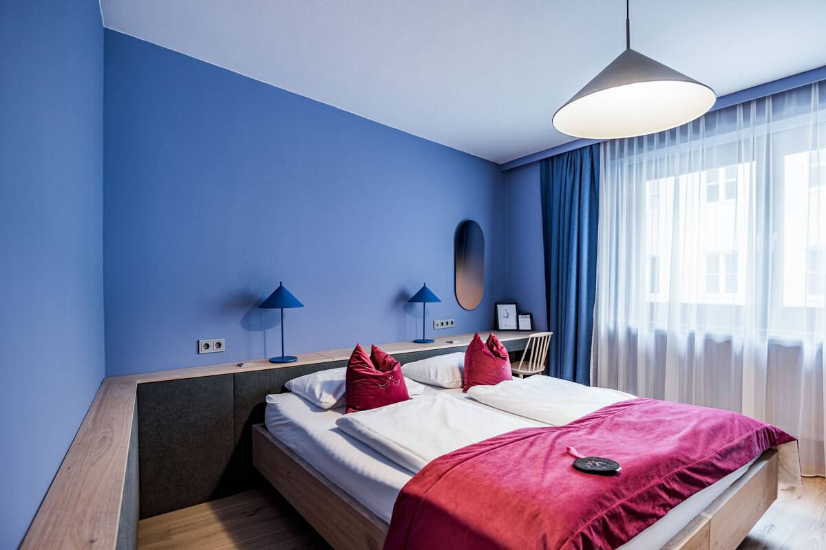 Una habitació d'hotel moderna amb un llit gran, dues làmpades de nit, un escriptori i una finestra amb cortines translúcides.