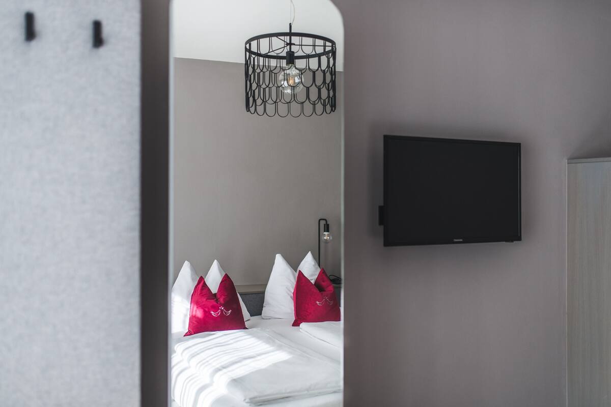 Habitació d'hotel moderna amb un llit, almoinades vermelles i blanques, un televisor negre fixat a la paret i una llum penjant negra i singular.