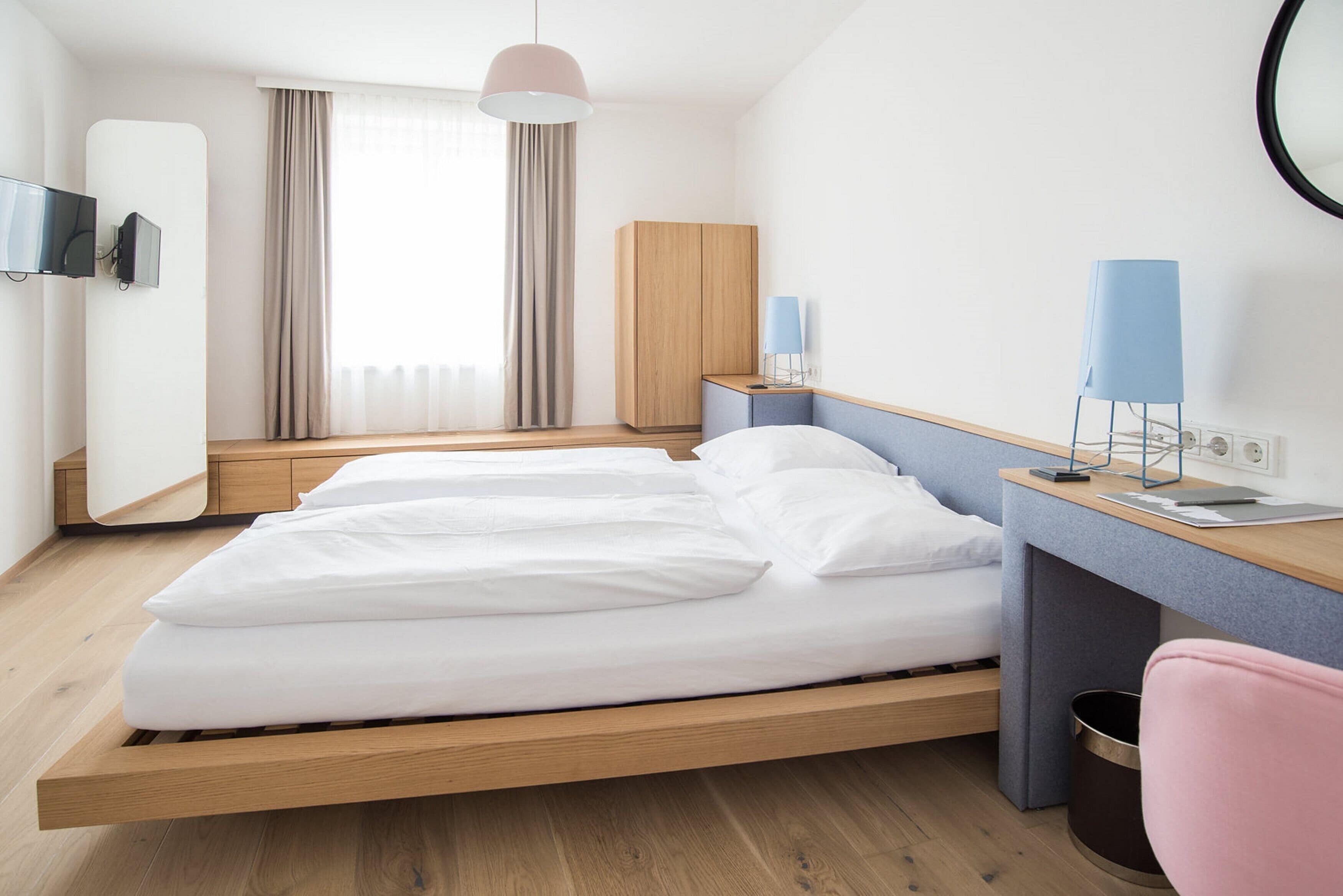 Ein modernes Hotelzimmer mit einem hölzernen Bett, einem Schreibtisch mit Lampe und Blick auf die Stadt durch große Fenster.