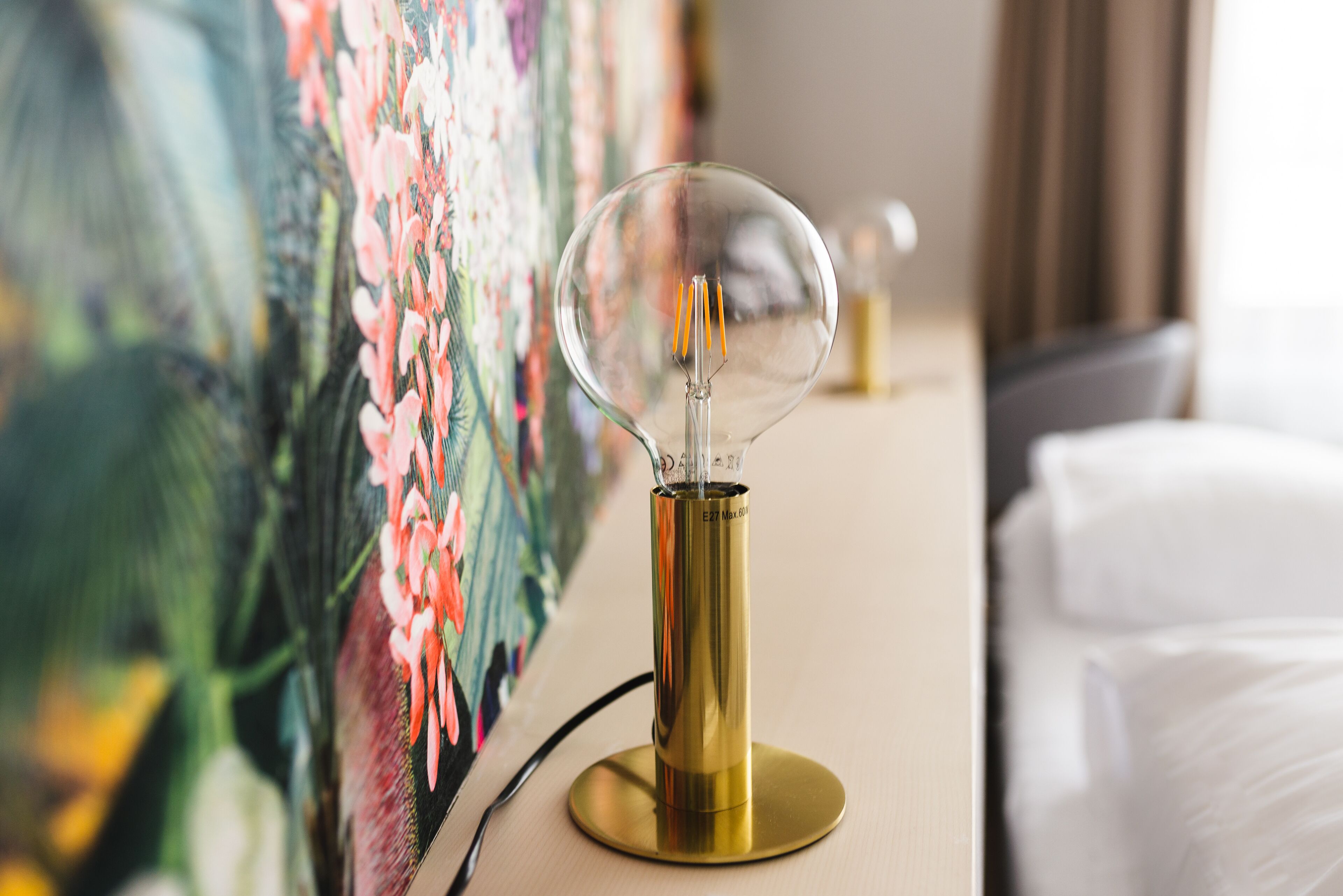Une lampe à ampoule en verre transparent, montée sur un pied en laiton, un tableau aux couleurs vives et un lit recouvert de draps blancs.