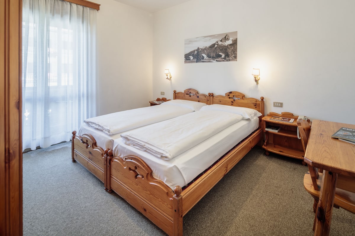 A double wooden bed with white linens, two bedside tables with lamps, a wooden desk, and a mountain landscape painting on the wall.