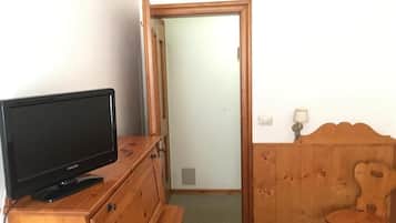 Une pièce comprenant une commode en bois, un téléviseur posé dessus, une chaise en bois et une porte donnant sur une autre pièce.
