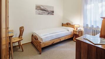 A bedroom with a wooden bed, a wooden desk, a chair, a nightstand with a lamp, and a window with sheer curtains.