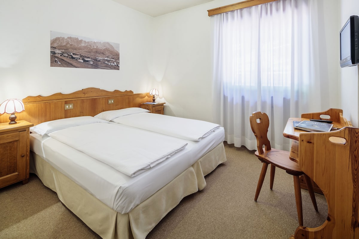 A hotel room with two beds, a desk with a chair, a lamp, a picture on the wall, and a window with curtains.