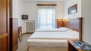 Habitació d'hotel amb un llit gran, un panell de capçalera de fusta, una taula de nit amb una revista, un televisor a la paret i una finestra amb cortines primes.