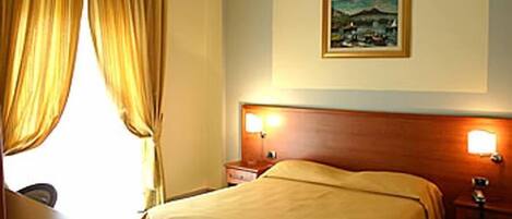 A hotel room with a bed, a nightstand, a lamp, a painting on the wall, and a window with curtains.