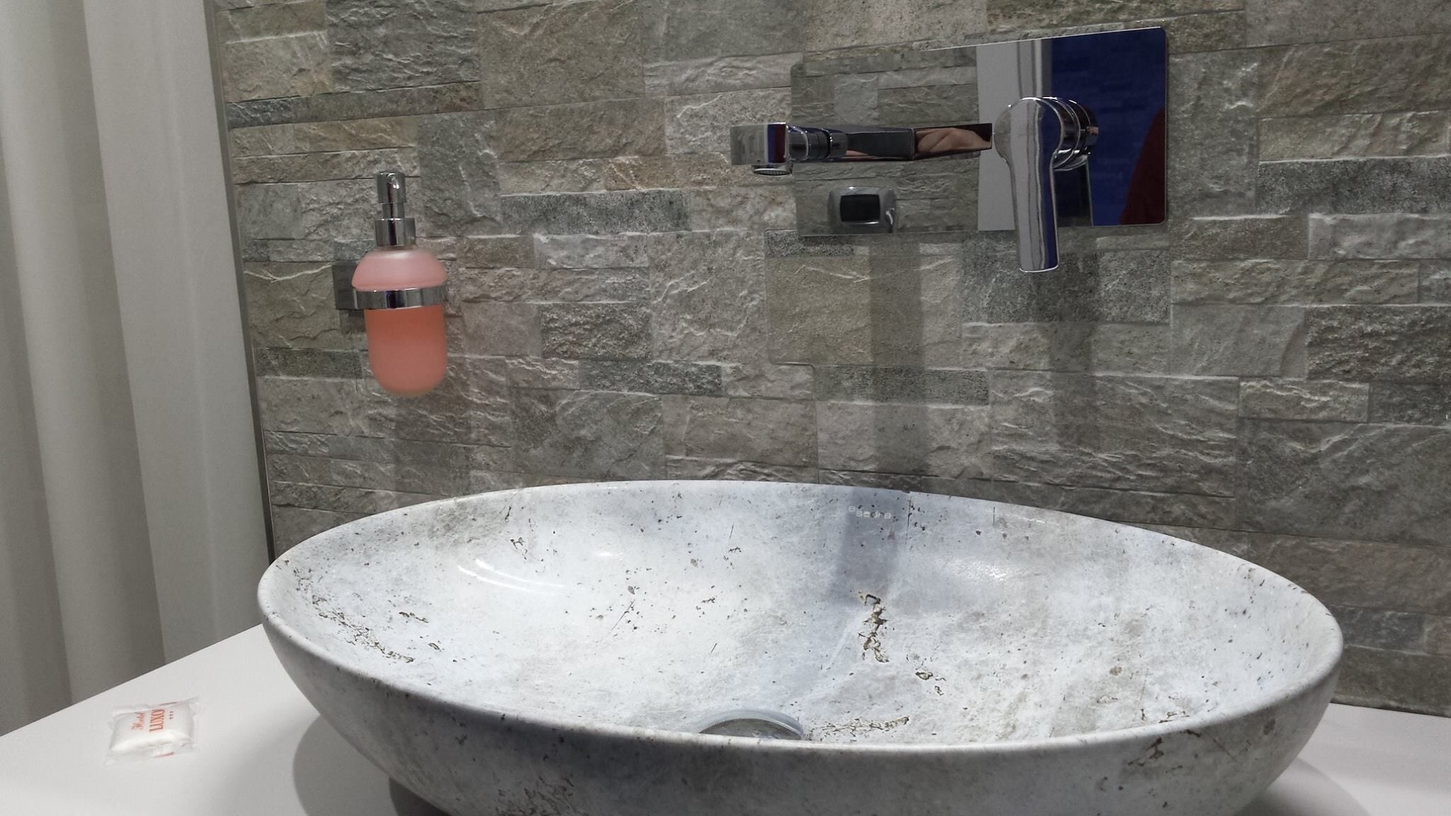Et badeværelse med en stor, plettet keramisk vask, en pink sæbedispenser og en moderne hane.