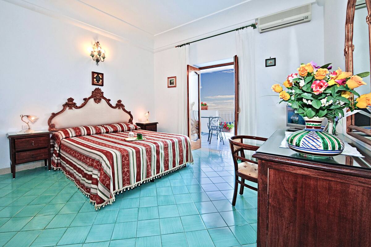 Une chambre à coucher avec un lit, un bureau, une chaise, un vase de fleurs et une porte-fenêtre donnant sur un balcon.