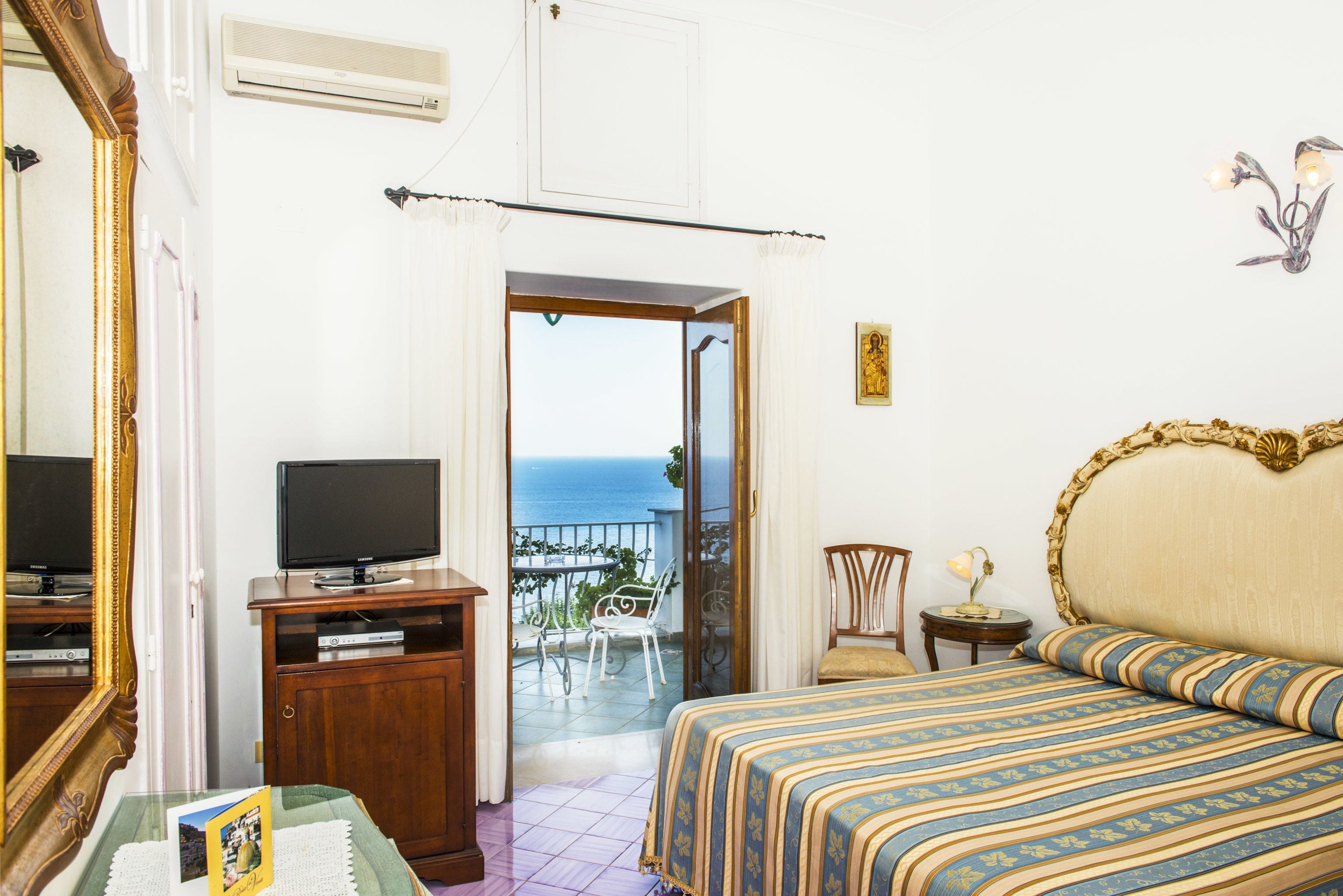 A bedroom with a bed, a television on a wooden cabinet, a small table with a lamp, and a view of the sea through an open door.