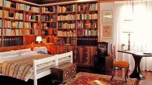 A cozy room with a wooden ceiling, a large bookshelf filled with books, a bed with a patterned bedspread, a small table with a lamp, and a chair. - At Home A Palazzo (Venice)