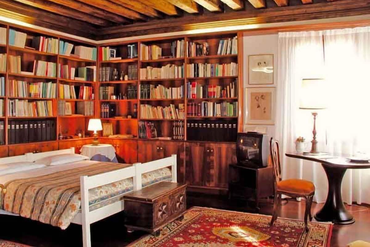 A cozy room with a wooden ceiling, a large bookshelf filled with books, a bed with a patterned bedspread, a small table with a lamp, and a chair.