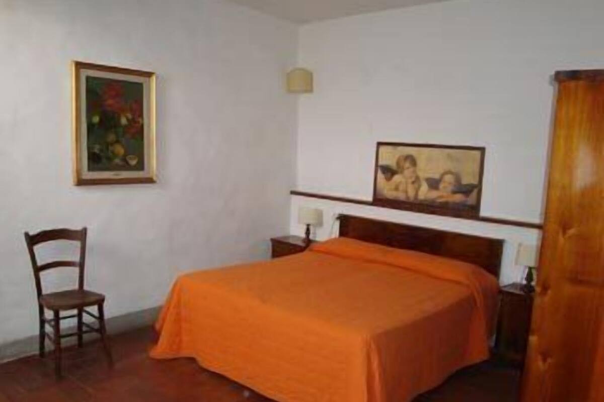 A bedroom with a wooden bed, an orange bedspread, a wooden chair, and two framed pictures on the wall.