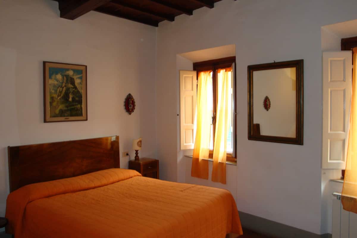 A bedroom with a wooden headboard, a bed with an orange bedspread, a nightstand, a framed picture on the wall, a window with curtains, and a mirror.