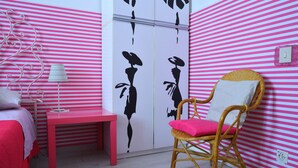 A room with a bed, a red table, a wicker chair with a white cushion, and a wall with pink and white stripes. - Leonardo's Rooms (Pontassieve)