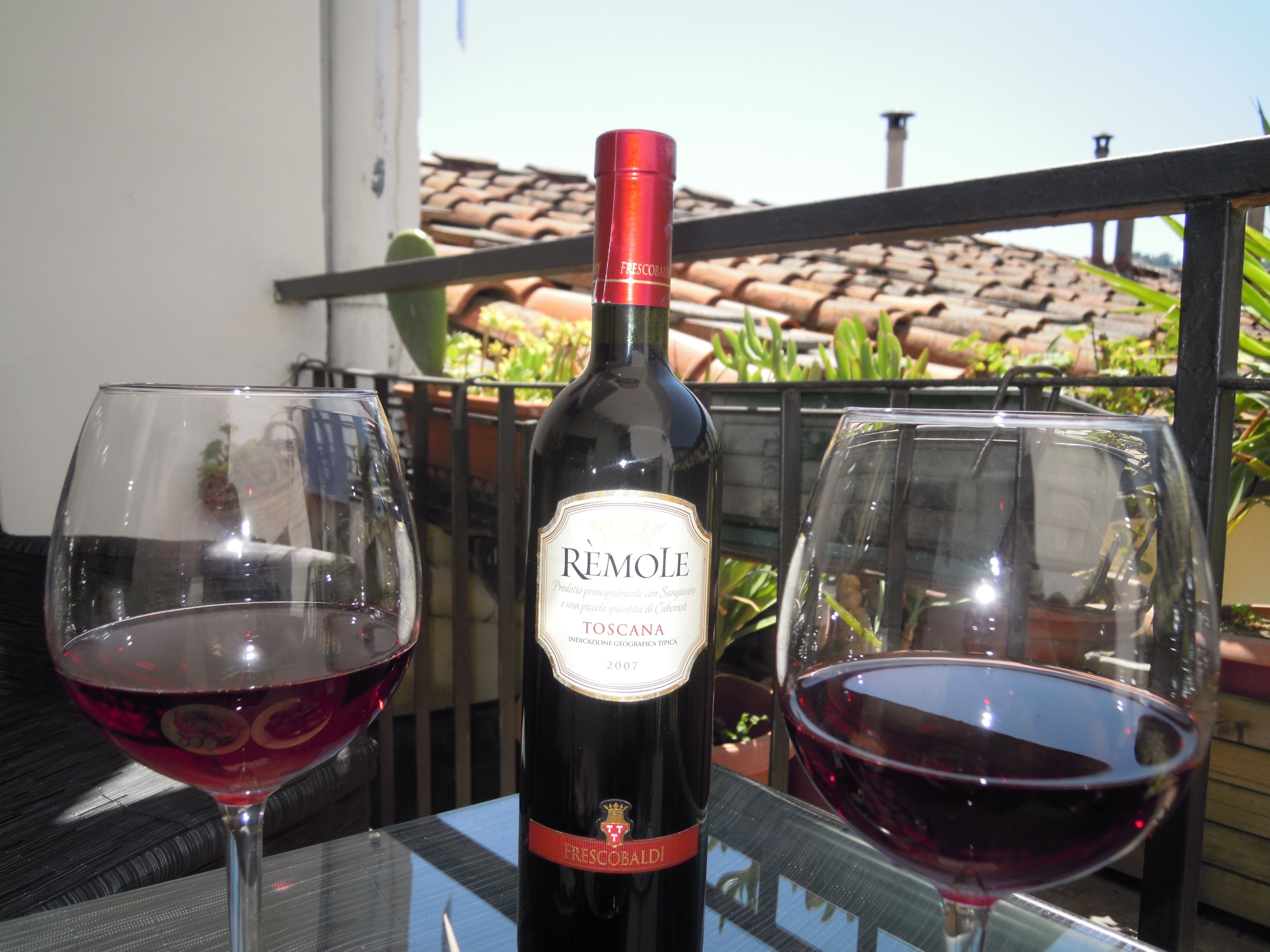 Une bouteille de vin Rèmole Toscana et deux verres de vin rouge sur un balcon doté d’un toit en tuiles et agrémenté, à l’arrière-plan, de plantes en pot.