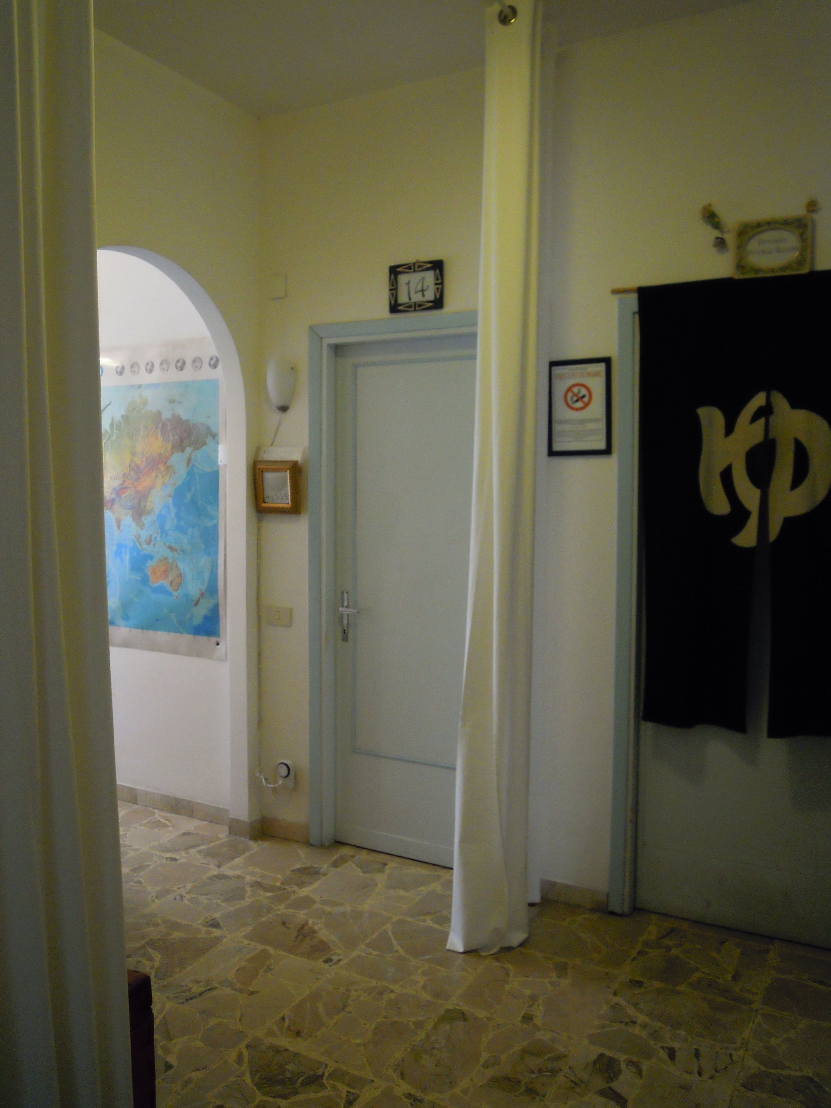 Un pasillo con piso de piedra, una puerta con número, un mapa en la pared y una toalla negra con un logotipo.