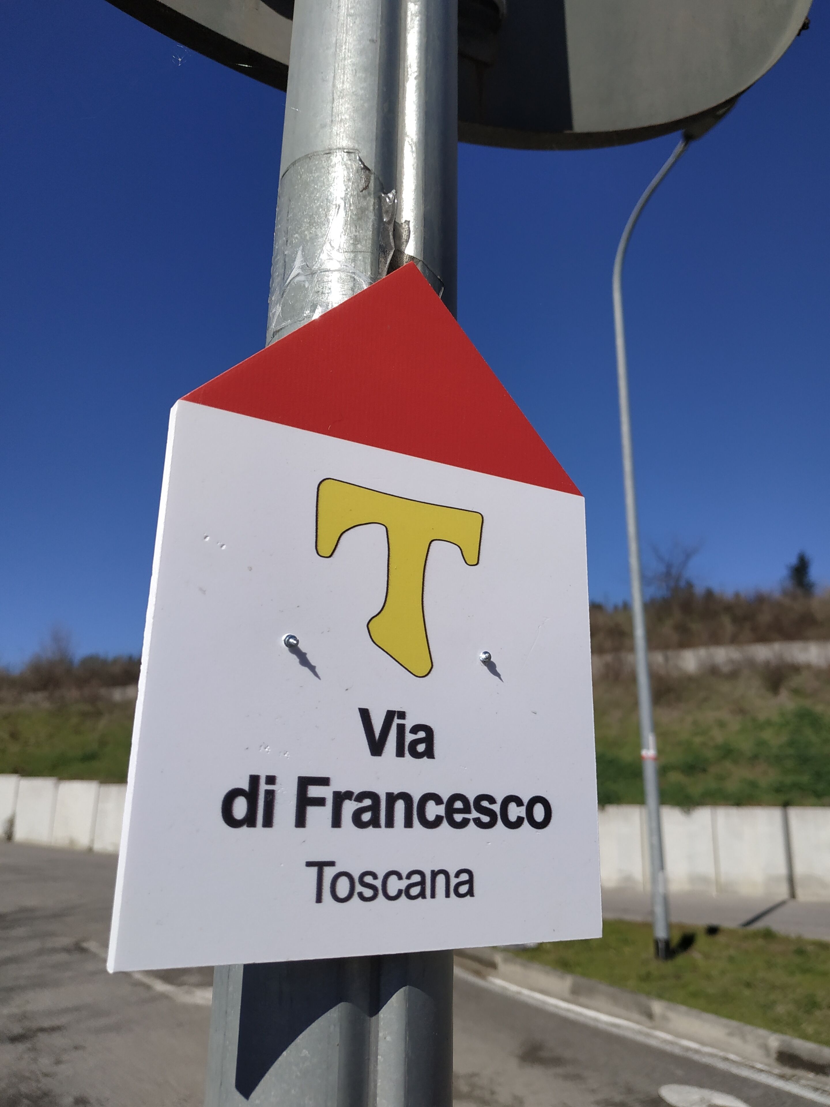 Señal con un triángulo rojo, una 'T' amarilla y el texto 'Via di Francesco Toscana'.