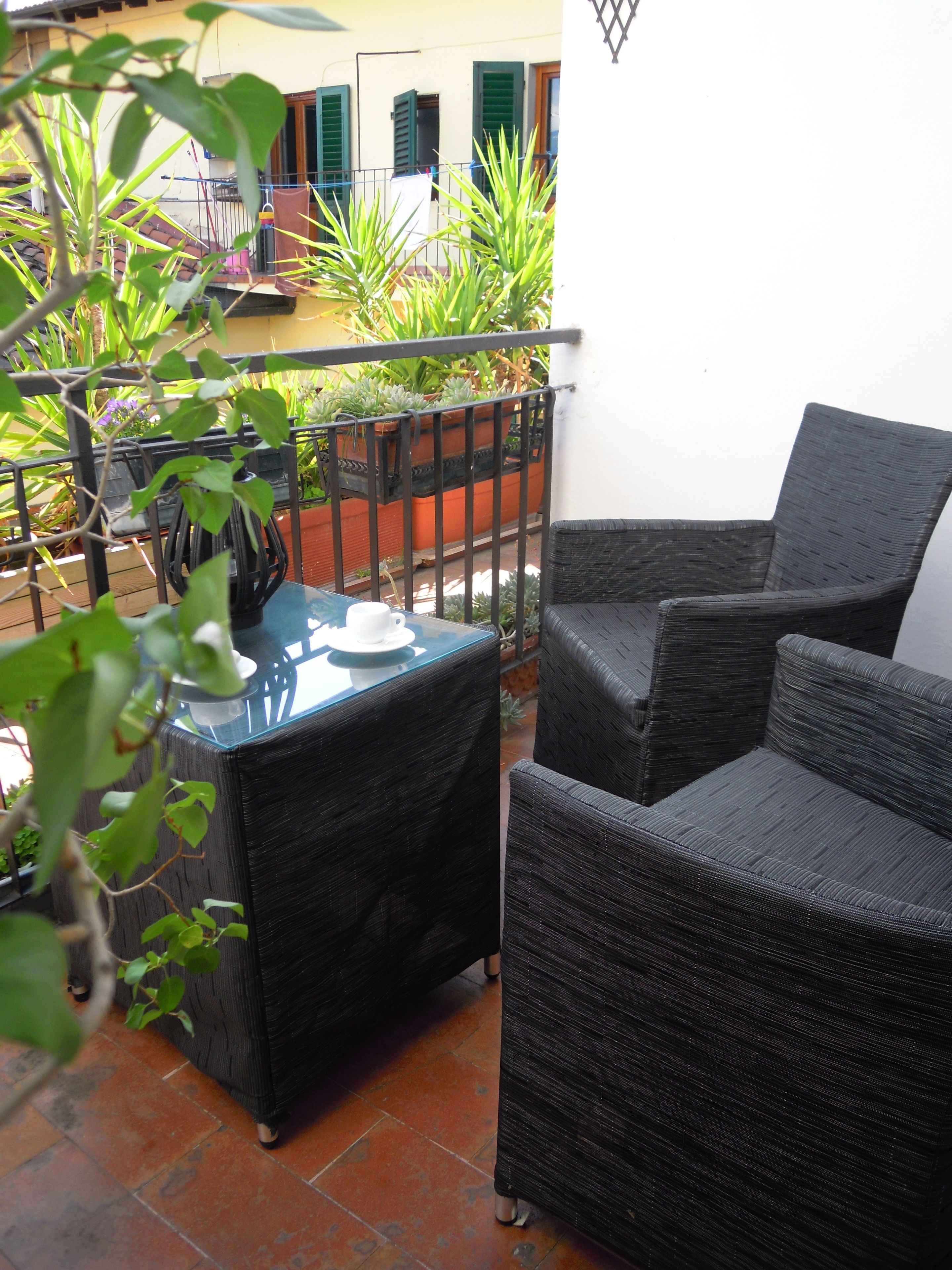 Un balcón con mobiliario de mimbre, una mesa de vidrio y plantas en macetas.