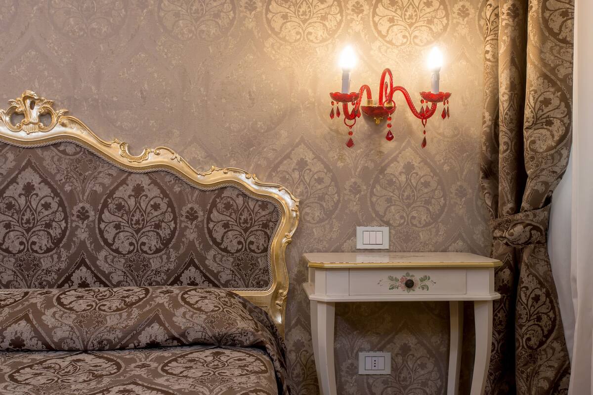 A bedroom with a patterned wallpaper, a gold-framed headboard, a white bedside table with a drawer, and a wall-mounted red candelabra.