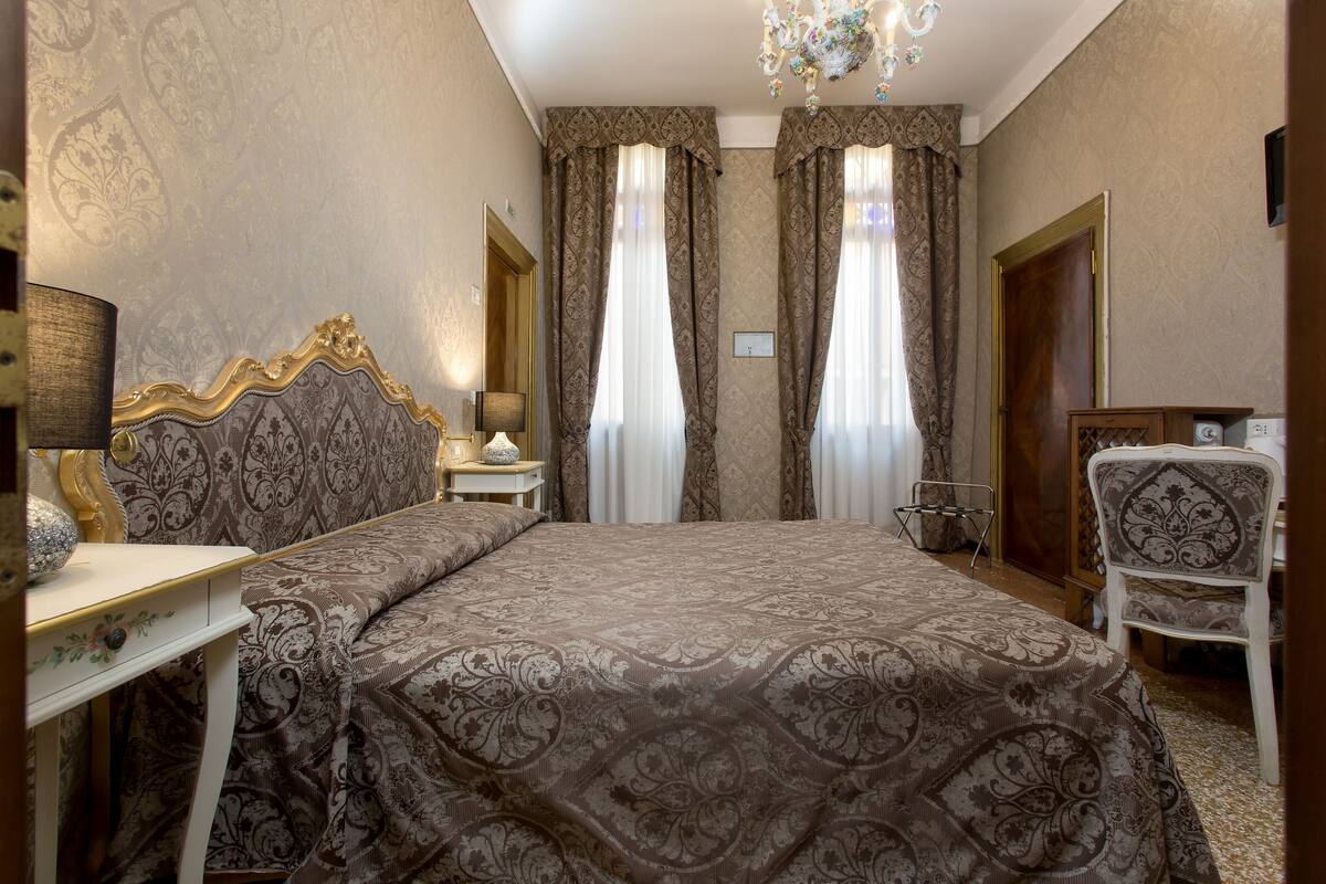 A hotel room with a large bed, a desk with a lamp, a chair, and a chandelier.