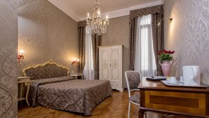 Ein Hotelzimmer mit einem Bett, einem Schreibtisch mit Stuhl, einem Kleiderschrank, einer Chandelüster und einer Vase mit Blumen.