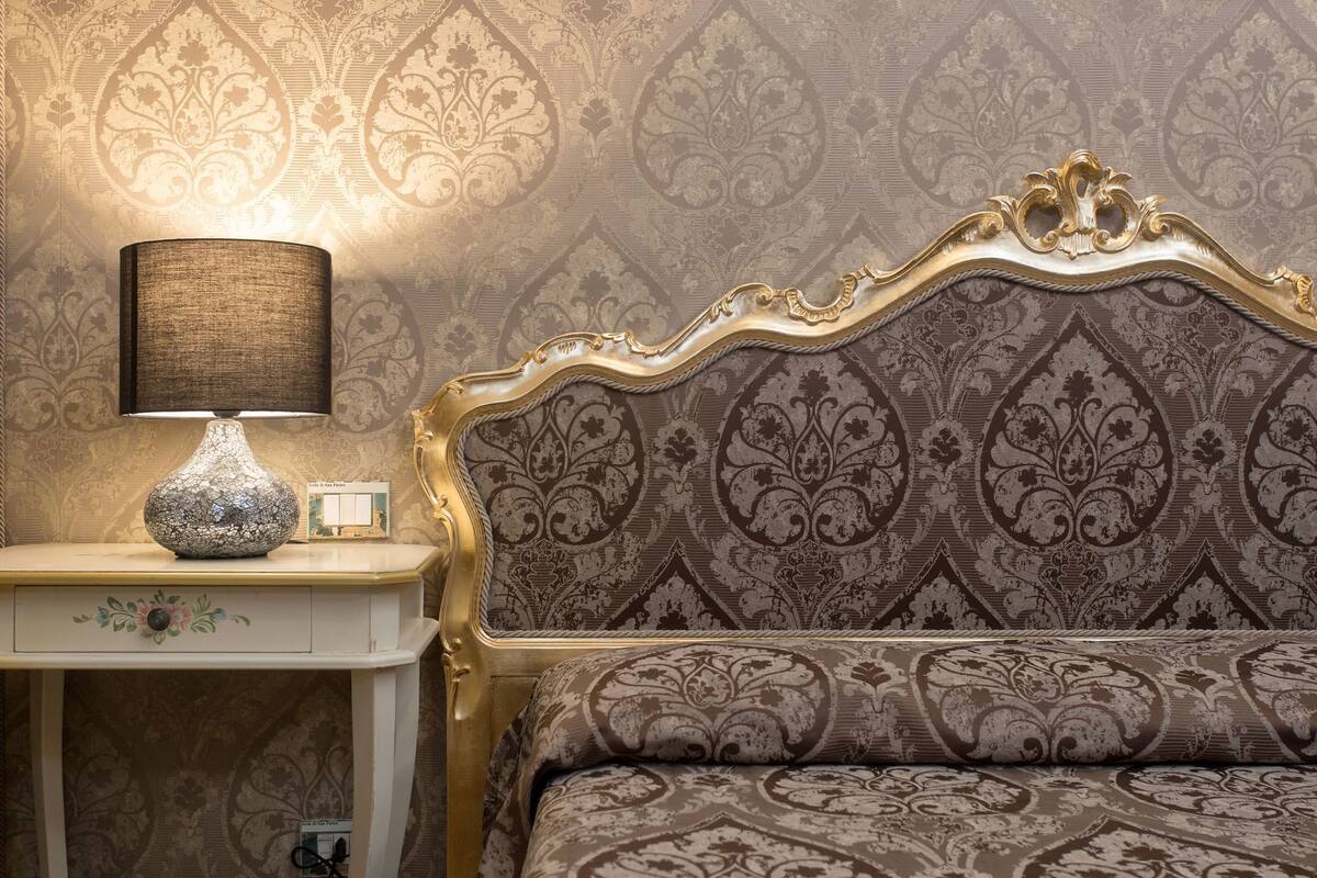 A bedroom with a patterned wallpaper, a decorative headboard, a bedside table with a lamp, and a small decorative item.