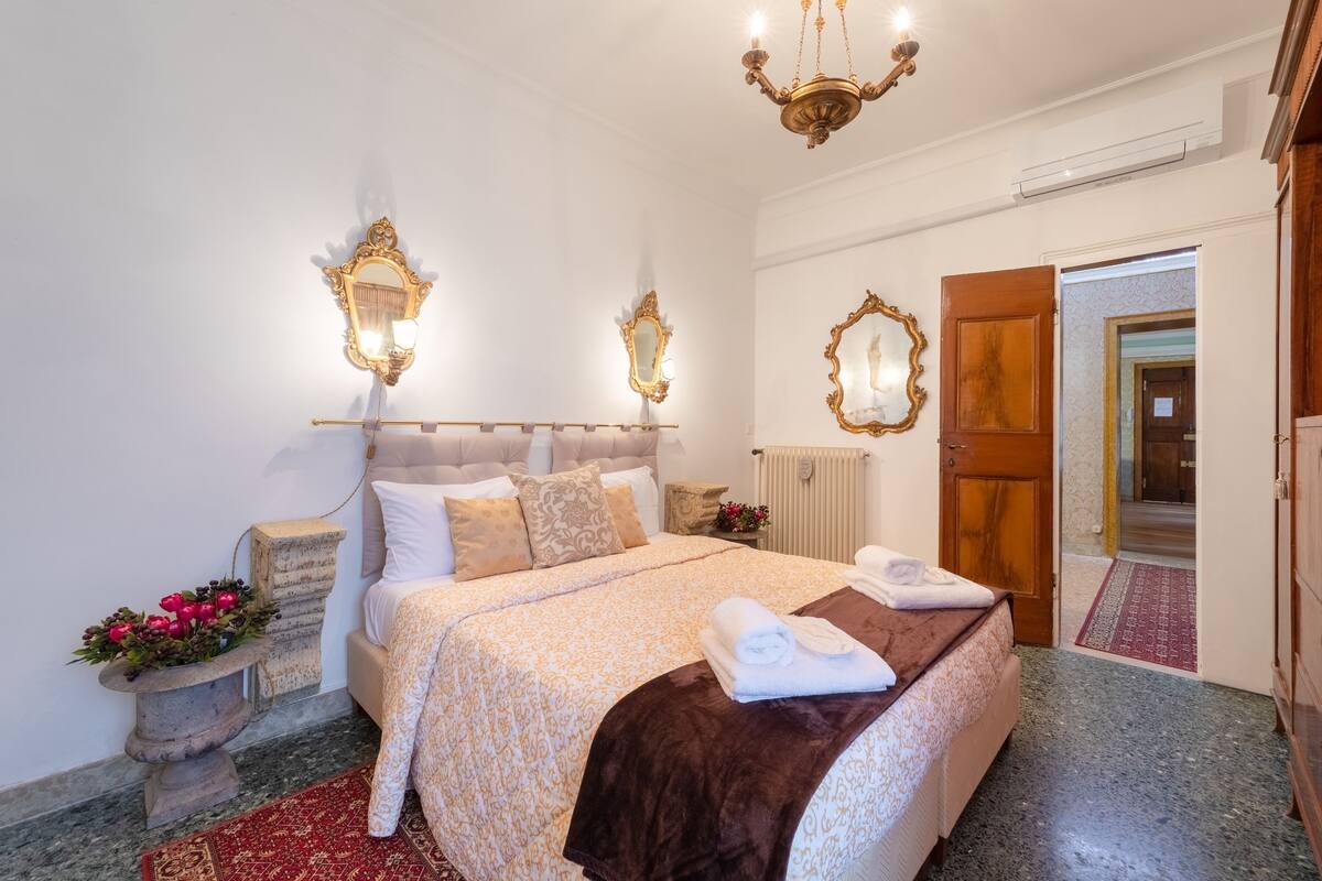 A well-lit bedroom with a large bed, two wall-mounted mirrors, a chandelier, and a wooden door.