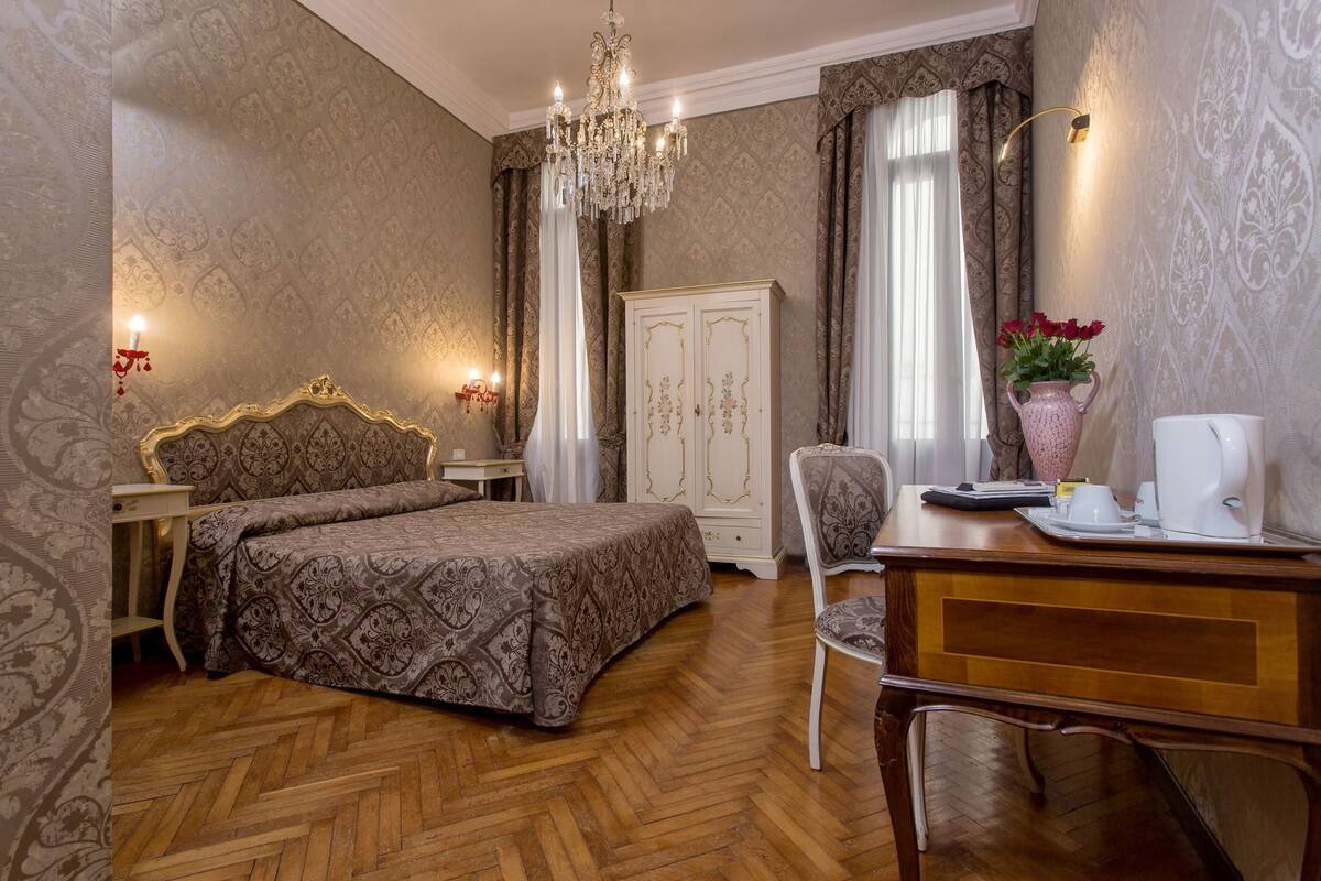 A bedroom with a bed, a desk, a chair, a wardrobe, a chandelier, and a vase with flowers.